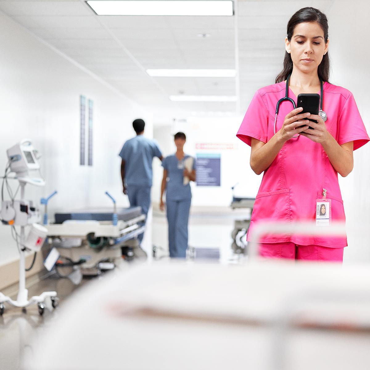 A clinician texts checks her phone while standing in a hospital corridor. She is wearing pink scrubs.