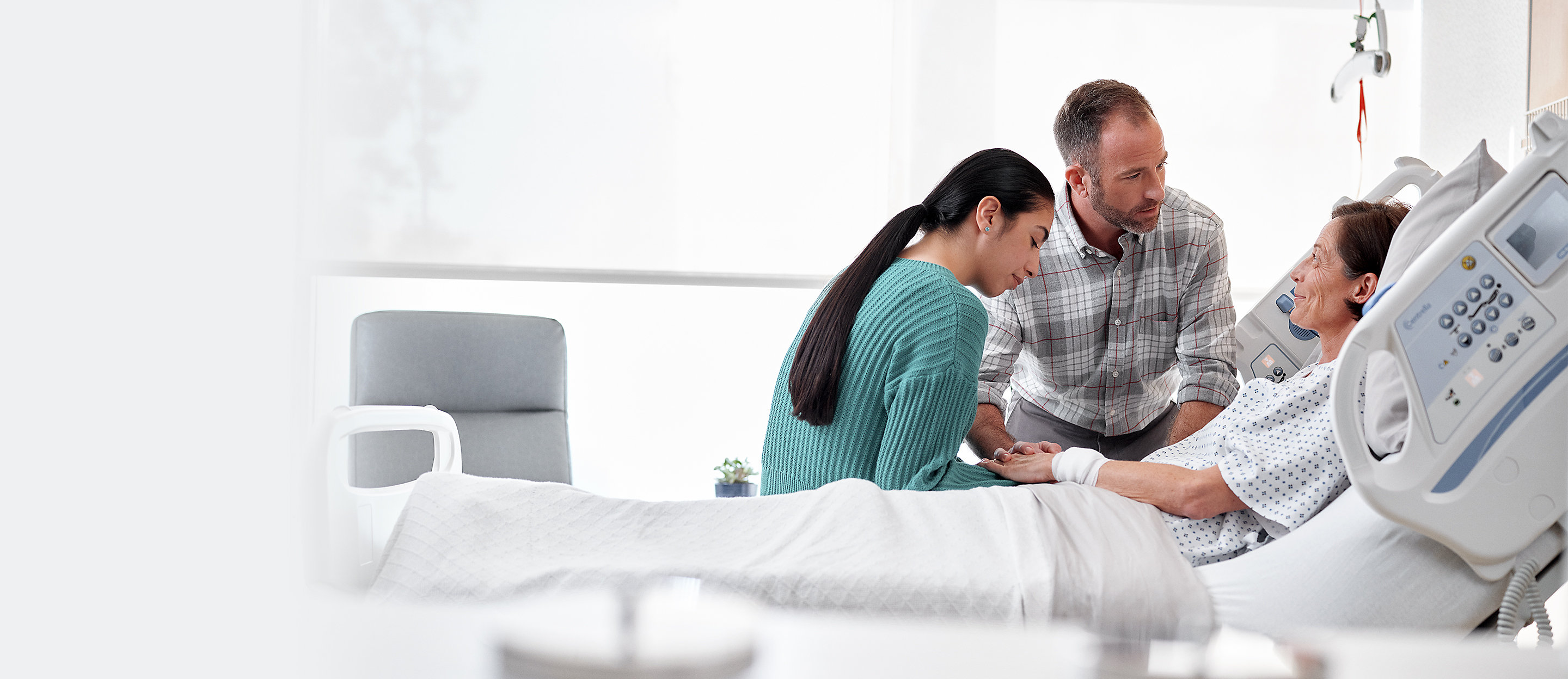 A man and woman hold the hand of a female patient who is in a hospital bed.